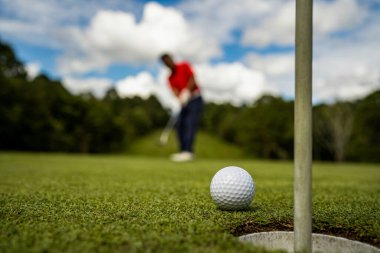 Golfçü topu yeşil golfün üstüne koyuyor, mercek akşam vakti parlıyor. Golfçü vuruşu uzun süre deliğe soktuktan sonra kazanır..
