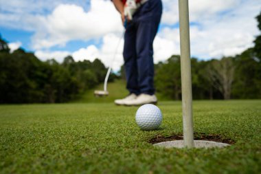 Golfçü topu yeşil golfün üstüne koyuyor, mercek akşam vakti parlıyor. Golfçü vuruşu uzun süre deliğe soktuktan sonra kazanır..