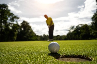 Golfçü topu yeşil golfün üstüne koyuyor, mercek akşam vakti parlıyor. Golfçü vuruşu uzun süre deliğe soktuktan sonra kazanır..
