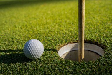 Odaklan. Golf deliğinin yanında beyaz bir golf topu ve yeşil çimlerin üzerinde bir bayrak direği güneşli arka planlar ve lens parlamaları için idealdir..