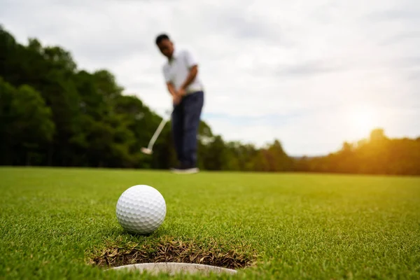 Golfçü topu yeşil golfün üstüne koyuyor, mercek akşam vakti parlıyor. Golfçü vuruşu uzun süre deliğe soktuktan sonra kazanır..