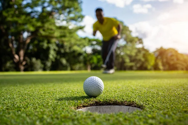 Golfçü topu yeşil golfün üstüne koyuyor, mercek akşam vakti parlıyor. Golfçü vuruşu uzun süre deliğe soktuktan sonra kazanır..