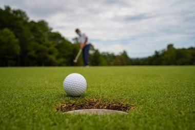 Golfçü topu yeşil golfün üstüne koyuyor, mercek akşam vakti parlıyor. Golfçü vuruşu uzun süre deliğe soktuktan sonra kazanır..
