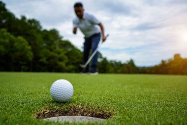 Golfçü topu yeşil golfün üstüne koyuyor, mercek akşam vakti parlıyor. Golfçü vuruşu uzun süre deliğe soktuktan sonra kazanır..