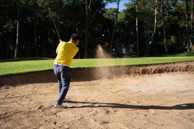 Asian golfer swings in a sand pit during pre-match practice at a golf course. A professional golfer hitting his ball out of a bunker with the sand and ball in mid-air.