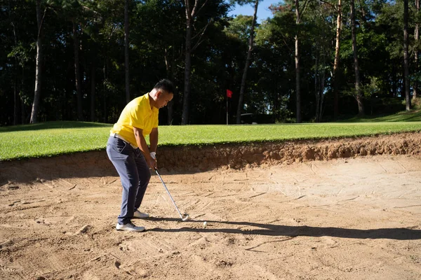 Asian golfer swings in a sand pit during pre-match practice at a golf course. A professional golfer hitting his ball out of a bunker with the sand and ball in mid-air.