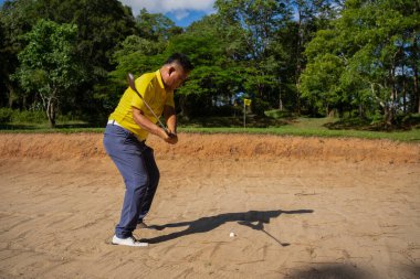 Asyalı golfçü bir golf sahasında maç öncesi antrenman sırasında kum havuzunda sallanıyor. Profesyonel bir golfçü topunu kumla sığınaktan dışarı atıyor..