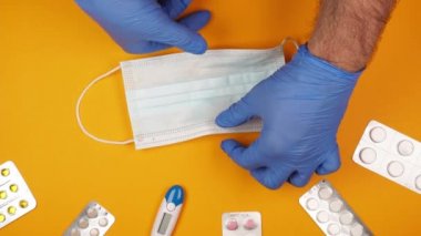 Mavi kauçuk eldivenli bir erkek doktorun elleri hastanın sıcaklığını ölçmek için tablet kabarcıkları ve termometreyle sarı arka planda tek kullanımlık bir maske tutuyor.