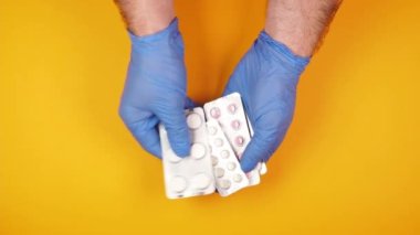 Mavi kauçuk eldivenli erkek doktorun ellerinde sarı arka planda bir sürü hap var.