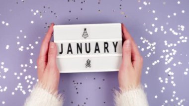 Beyaz kabarık kazaklı bir kadın elleri üzerinde Ocak ayının mor arka planında parıldayan altın yıldızlarla birlikte beyaz bir tahta tutuyor.