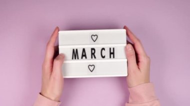 Genç bir kızın elleri üzerinde pembe arka planda kalpler olan beyaz bir tahta tutuyor.