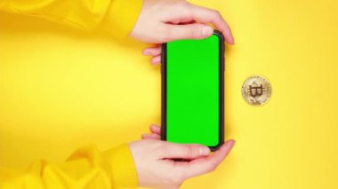 Dikey video Genç kadın tüccar ve yatırımcı ellerinde yeşil krom anahtarlı akıllı telefon altın parlayan bitcoin ile parlak sarı arka planda takas uygulaması