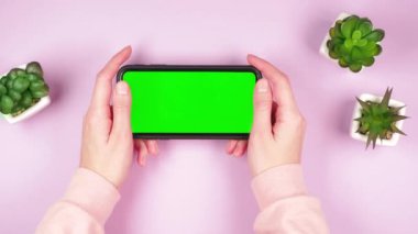 Elinde yeşil renkli akıllı bir telefon tutan genç bir kızın elleri. Pembe arka plandaki uygulamalar için üç yeşil iç mekan bitkisi.