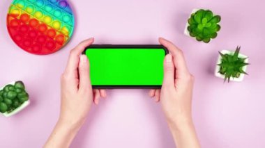 Genç kadın, elinde yeşil krom anahtar uygulamalı akıllı telefon, pembe arka planda, gökkuşağı silikon oyuncak ve yeşil bitkilerle birlikte.