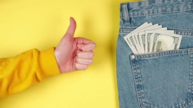 Parlak sarı arkaplanlı gençlerin cep harçlığı üzerinde kot pantolon cebinde bir sürü doların olduğu arka planda baş parmak gösteren bir kadın el.