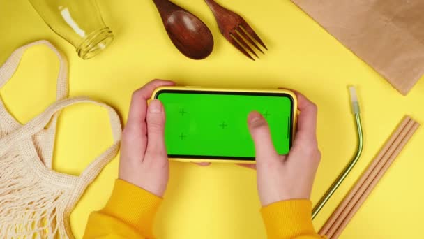ショッピングバッグ 環境に優しい木製フォークやスプーン ガラス瓶や再利用可能なストローと黄色の背景に緑の画面クロマキーアプリのモックアップとスマートフォンを保持若い女性の手 — ストック動画