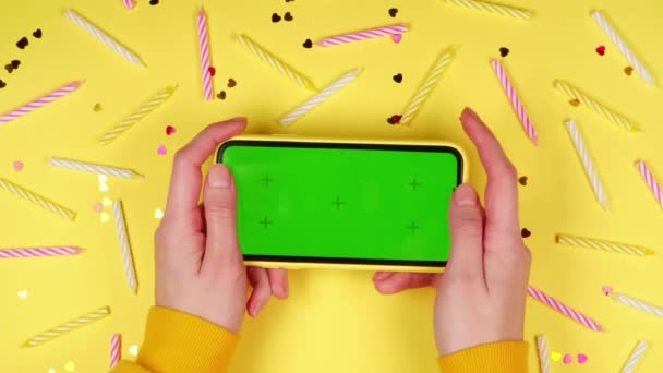 緑の画面でスマートフォンを保持している若い女性の手Chromakeyアプリは 多くのカラフルな誕生日ケーキキャンドルと黄色の背景にモックアップ — ストック動画