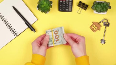 Genç bir kızın elleri, hesap makinesi, gayrimenkul anahtarları, ev bitkileri ve kalemli bir not defteri olan parlak sarı bir zemin üzerinde yüzlerce dolarlık banknotlar sayıyor.