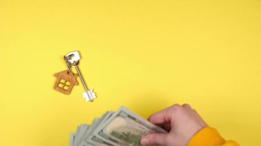Genç bir kadının eli, sarı arka plandaki yeni bir mülkten alınmış ahşap bir anahtarlıktaki gümüş bir anahtarlığın yanına bir sürü 100 dolarlık banknot koyar.