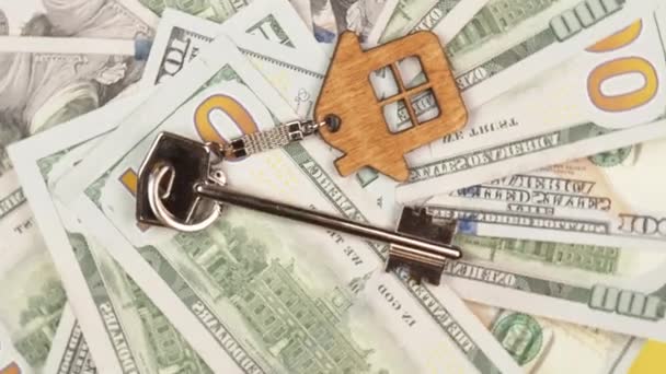 在几百美元钞票的背景下 一把装有木制钥匙链的银色钥匙正在旋转 钥匙的形式是新屋的房子 — 图库视频影像