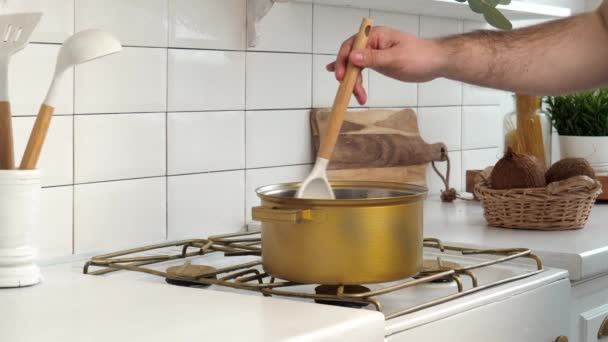 在一个白色的现代化厨房里 一个年轻人的手在关掉的煤气炉上 用金色平底锅搅拌着汤 — 图库视频影像
