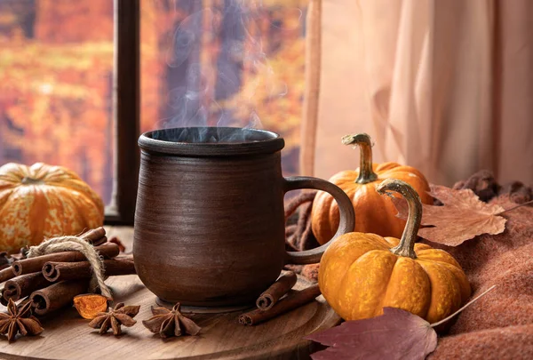 Renkli bir sonbahar arka planına sahip pencerenin yanında dumanı tüten sıcak çikolata ve mini balkabakları.