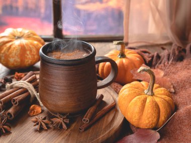 Renkli bir sonbahar arka planına sahip pencerenin yanında dumanı tüten sıcak çikolata ve mini balkabakları.