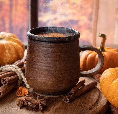 Renkli bir sonbahar arka planına sahip pencerenin yanında dumanı tüten bir bardak sıcak çikolata ve mini balkabağı.