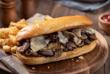 Biftek, peynir ve soğanla yapılmış Philly peynirli sandviç. Tahtada patates kızartması ve kızarmış sandviç.