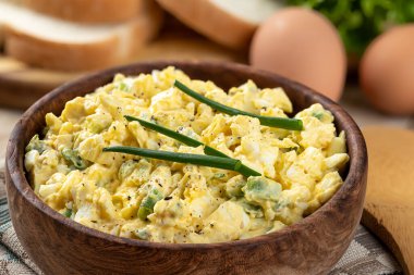 Arka planda yumurta, ekmek ve marul olan ahşap bir kasede yumurta salatası.