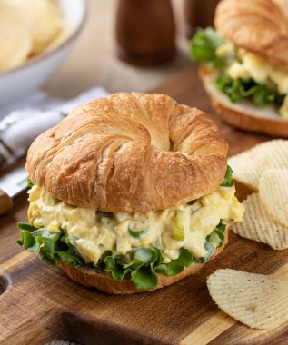 Kruvasan üzerinde marullu yumurta salatalı sandviç ve tahta kesme tahtasındaki patates cipsi.