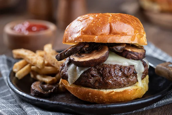 Closeup of a mushroom cheeseburger on a toasted bum with french fries on a black plate