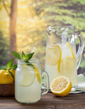 Limonata, limon dilimleri ve bardakta naneli sürahi ve arka planda limonlu tahta masa ve kırsal arka plan.