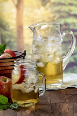 Elma dilimi ve buzlu bir bardak elma suyu ve bir sepet elma ve sürahi kırsal yaz arka planına sahip ahşap bir masada.