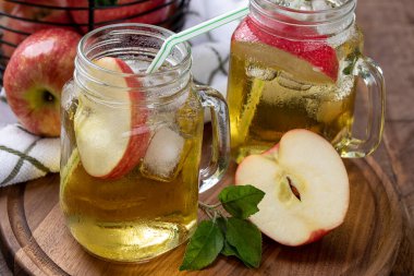 İki bardak elma suyu, elma dilimleri ve arka planda elma sepeti olan ahşap bir tabakta buz.