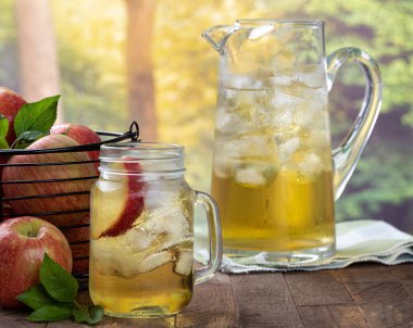 Elma dilimi ve buzlu bir bardak elma suyu ve bir sepet elma ve sürahi kırsal yaz arka planına sahip ahşap bir masada.