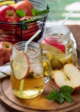 İki bardak elma suyu, elma dilimleri ve arka planda elma sepeti olan ahşap bir tabakta buz.