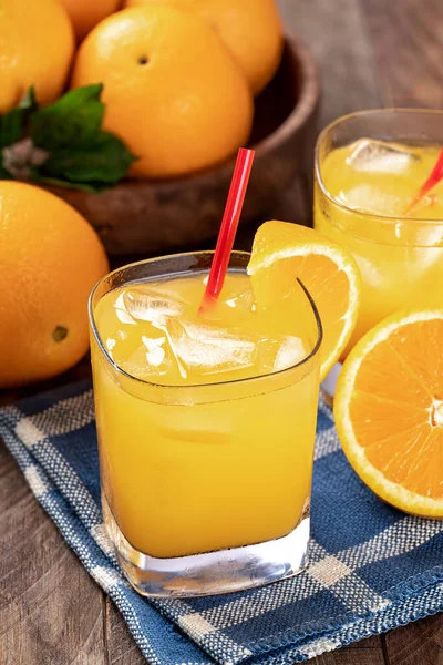 Glass of cold orange juice and sliced oranges on a wooden table with bowl of oranges in background