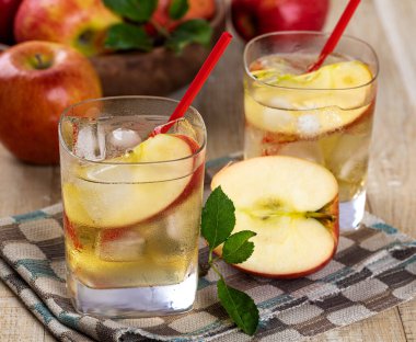 Arka planda dilimlenmiş elma ve elmalar olan soğuk bir bardak elma suyu.