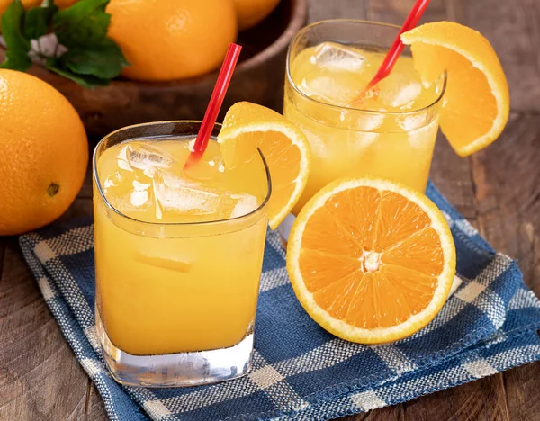 Glass of cold orange juice and sliced oranges on a wooden table with bowl of oranges in background