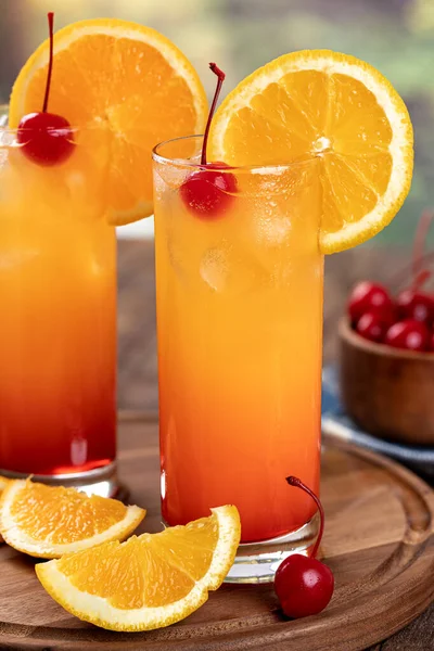 Two tequila sunrise cocktails with orange slice and cherry on a rustic wooden platter
