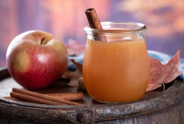 Glass of apple cider with cinnamon sticks and an apple on a wooden barrel with autumn background