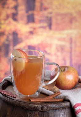 Glass cup of apple cider with apple slice on a wooden barrel with colorful autumn background