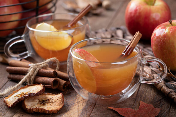 Cup of apple cider with cinnamon stick and sliced apple on a wooden table with fresh apples in background