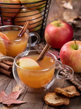 Cup of apple cider with cinnamon stick and sliced apple on a wooden table with fresh apples in background