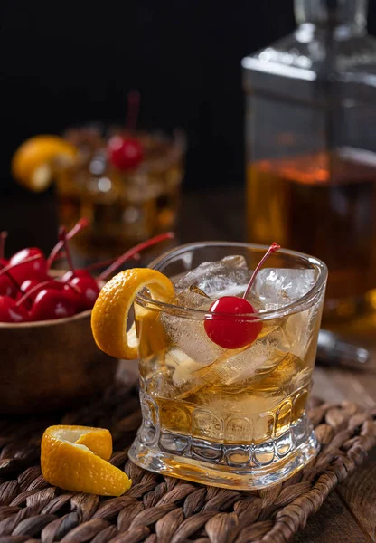 Old fashioned cocktail with orange peel and cherry on a wooden table with bowl of cherries and whiskey bottle