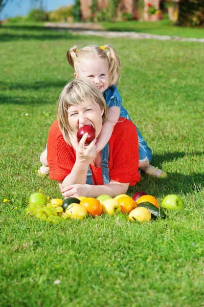 Matka a dcera jíst barevné ovoce na zelené trávě — Stock fotografie