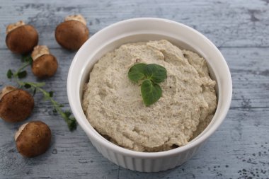 Mushroom Pate in a white bowl, on a blue surface, close up view, topped with fresh basil, brown champignon mushrooms on the side