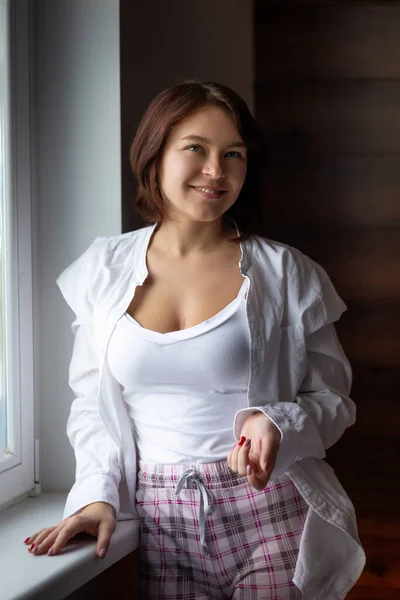 Güzel esmer kadın pencerede gömlek ve pijama giyer, fotoğraf pozu verir, poz verir, modellik, mankenlik, depresyon, depresyon, bezginlik, sabah ritüelleri eğitimi alır.