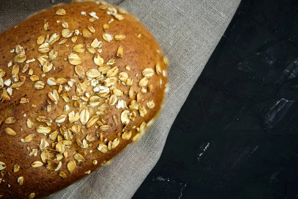 Rye bread sprinkled with oatmeal on a background of gray linen fabric and a black table. Loaf of bread top view. Sourdough bread close up with place for text